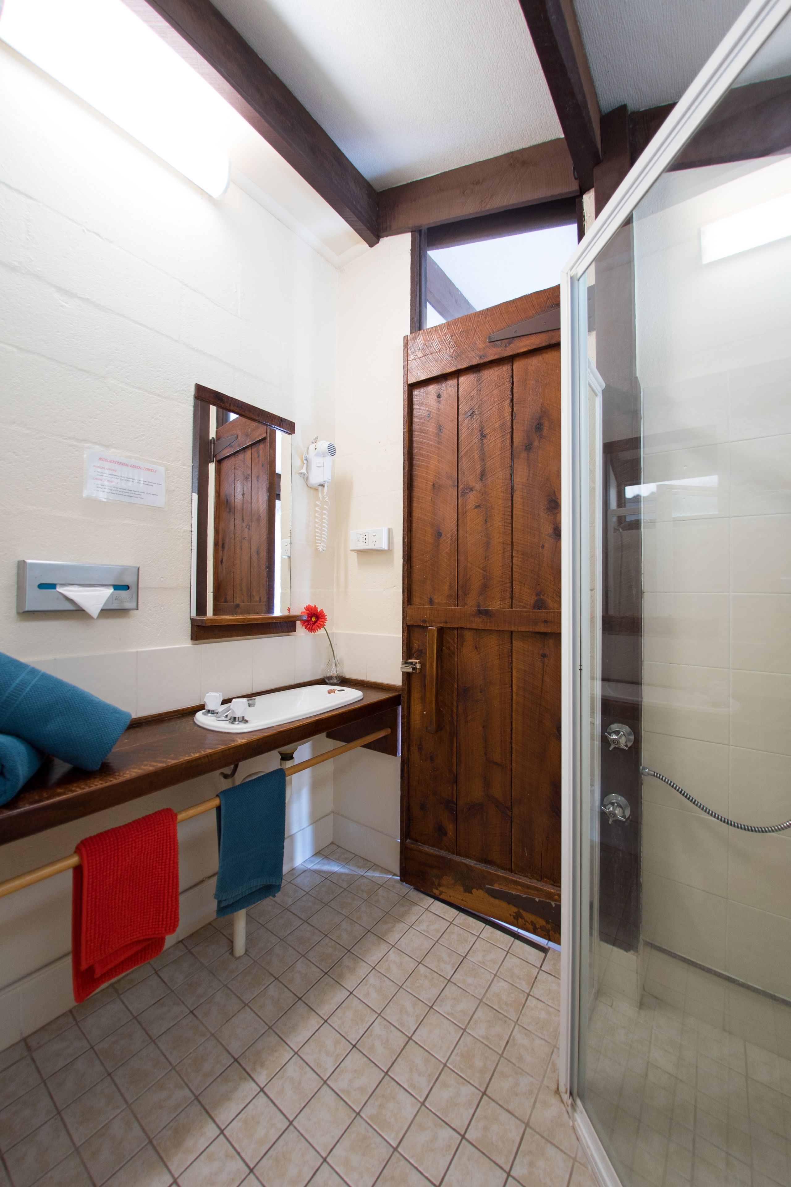 A bathroom with a wooden door, a sink with a mirror, a towel rack with red and blue towels, and a shower area.