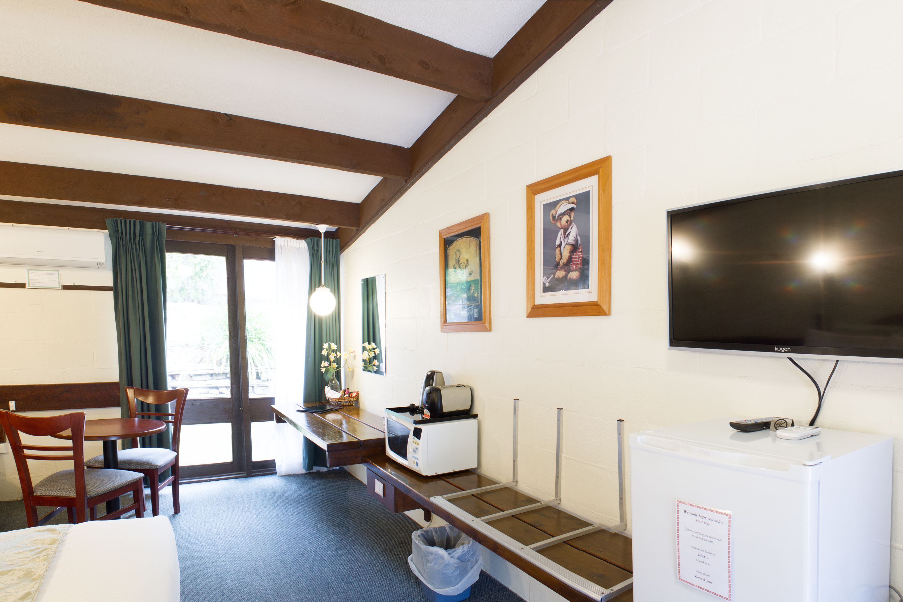 Habitación de hotel con cama, escritorio con computadora, televisor y refrigerador pequeño. Hay dos sillas y una ventana con cortinas.
