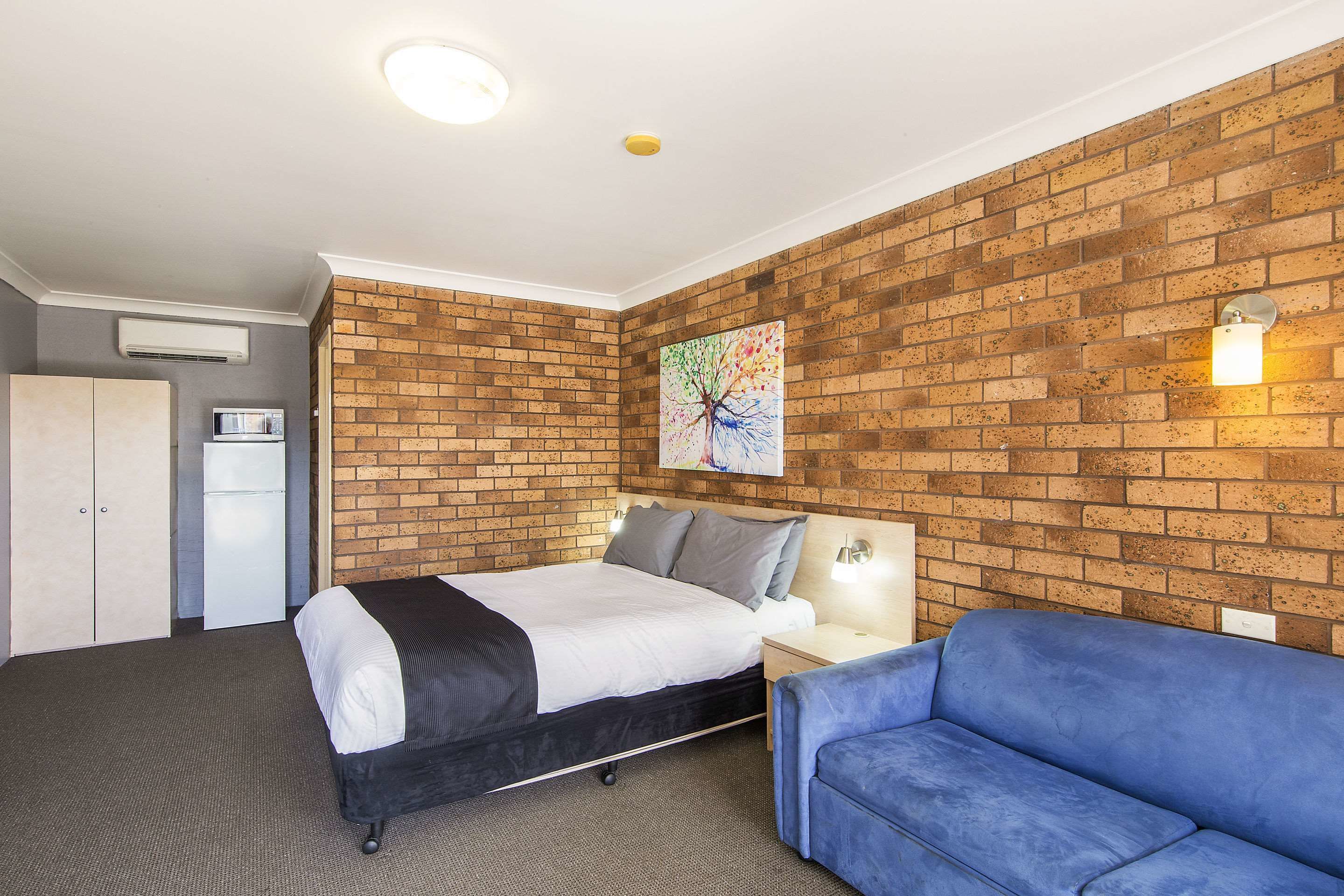 A hotel room with a brick wall, a bed, a blue sofa, a nightstand, and a painting.