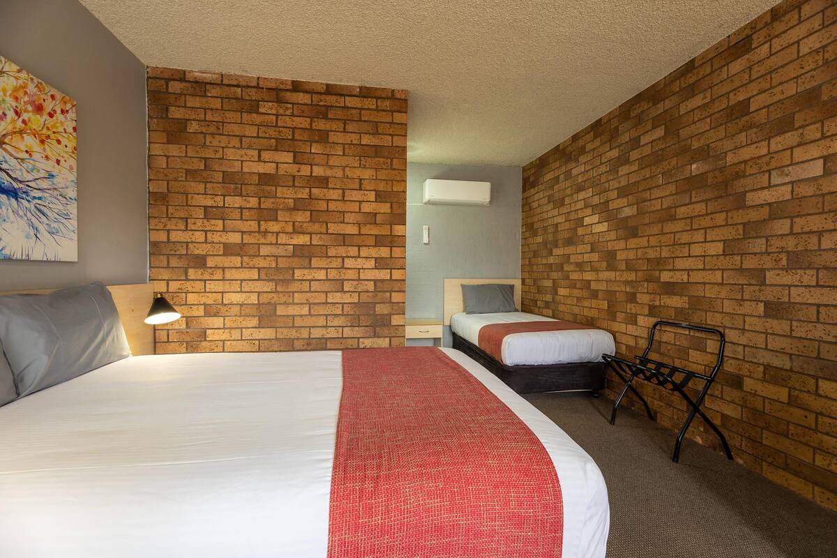 Una habitación de hotel con pared de ladrillo, una cama grande con una manta roja, una cama más pequeña, un escritorio con una silla y un cuadro en la pared.