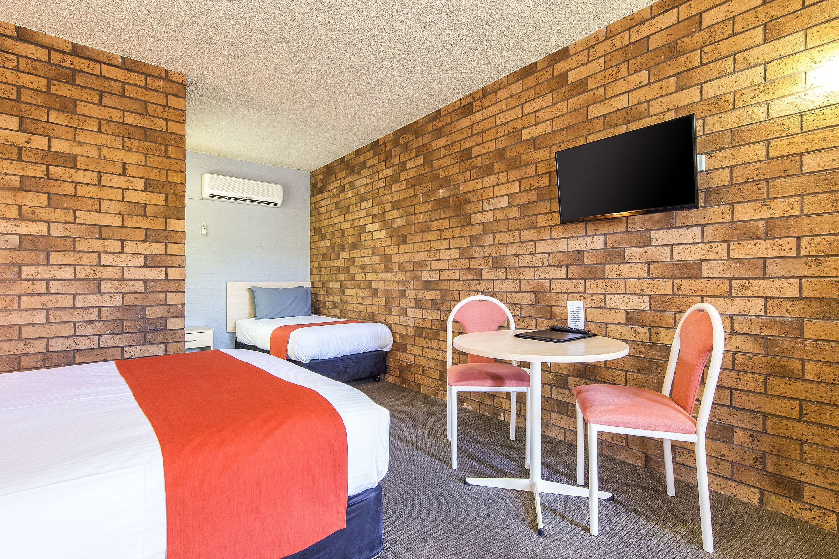 A hotel room with two beds, a small table with chairs, a flat-screen TV mounted on the brick wall, and an air conditioning unit.