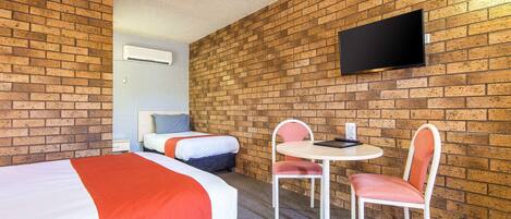 A hotel room with two beds, a small table with chairs, a flat-screen TV mounted on the brick wall, and an air conditioning unit.