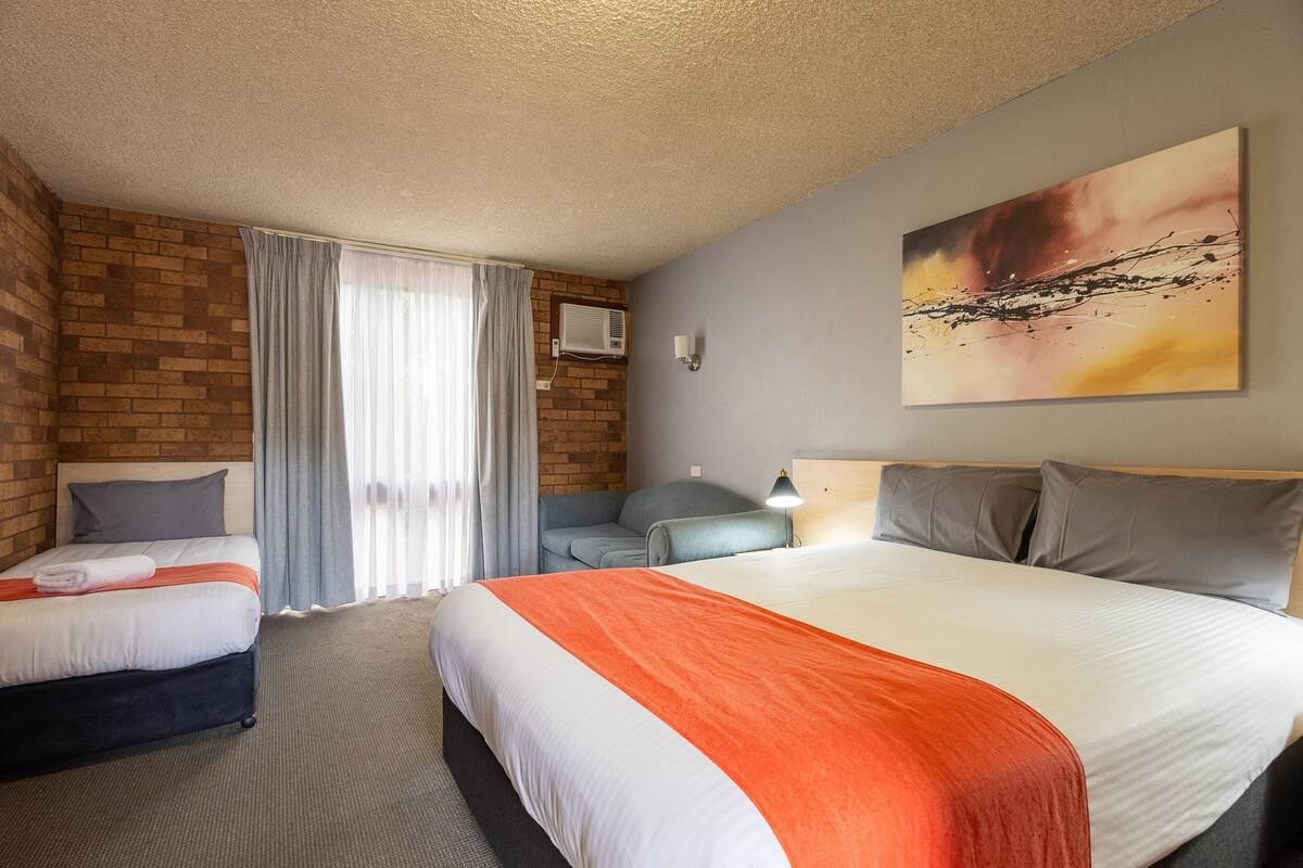Habitación de hotel con una cama grande, una cama más pequeña, un escritorio y una silla. Se ve una ventana con cortinas, un cuadro en la pared y un equipo de aire acondicionado.