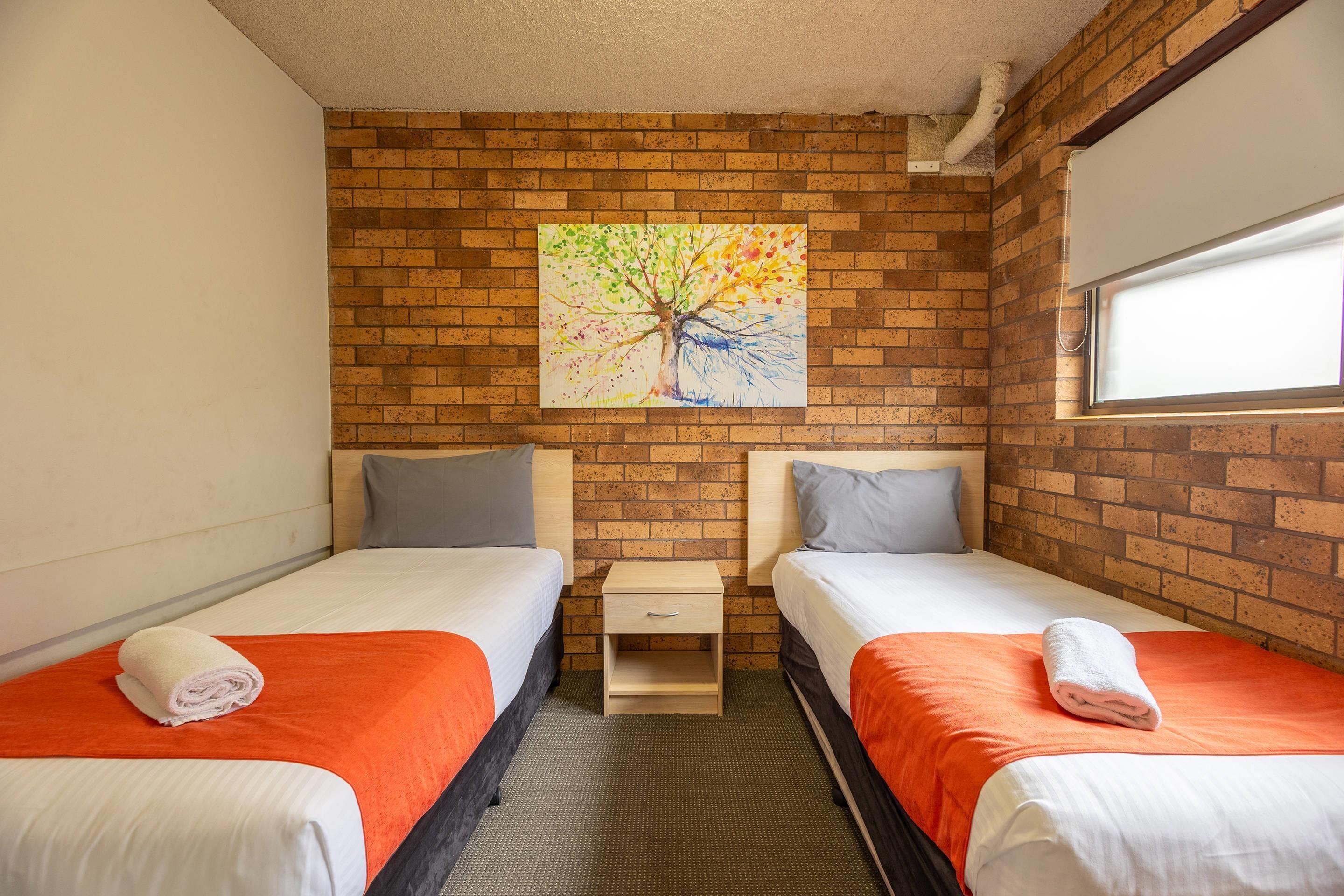 A hotel room with two beds, a brick wall, a painting of a tree, and a window.