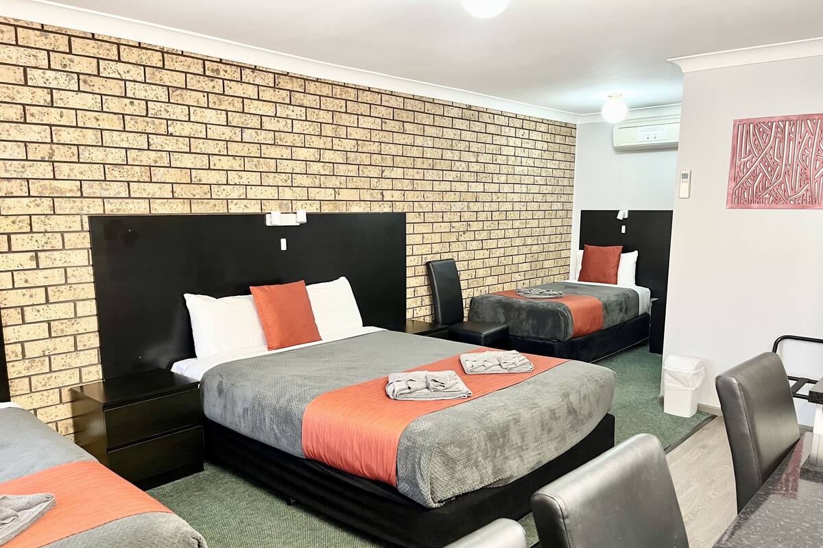 Una habitación de hotel con pared de ladrillo, dos camas con cabeceros negros, ropa de cama gris y naranja, una mesita y una silla.