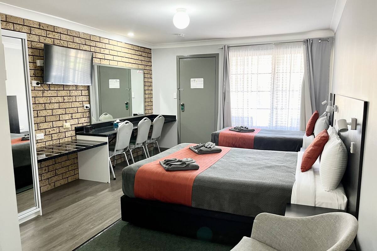 Habitación de hotel con dos camas, un escritorio con sillas, un televisor y una pared de ladrillo.