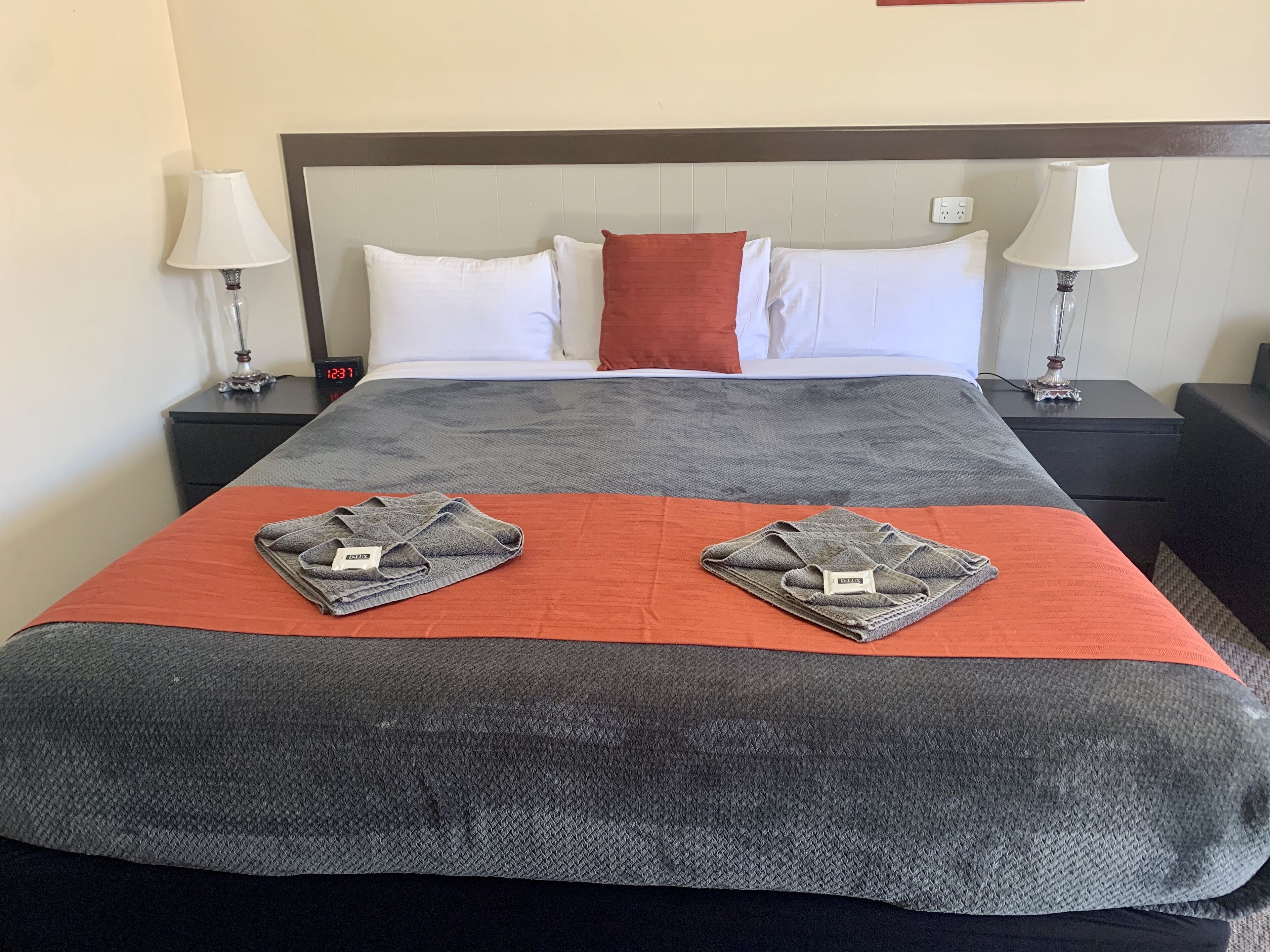 A neatly made bed with a grey and orange bedspread, two folded towels on the bed, white and red pillows, and two bedside lamps on nightstands.