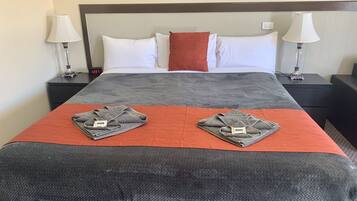 A neatly made bed with a grey and orange bedspread, two folded towels on the bed, white and red pillows, and two bedside lamps on nightstands.