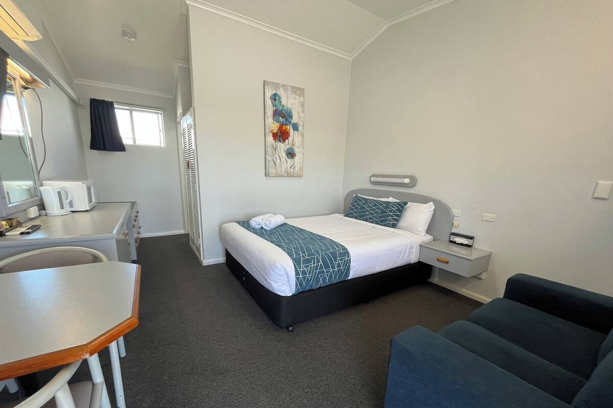A compact hotel room with a bed, bedside table, chair, and a small desk with a kettle.