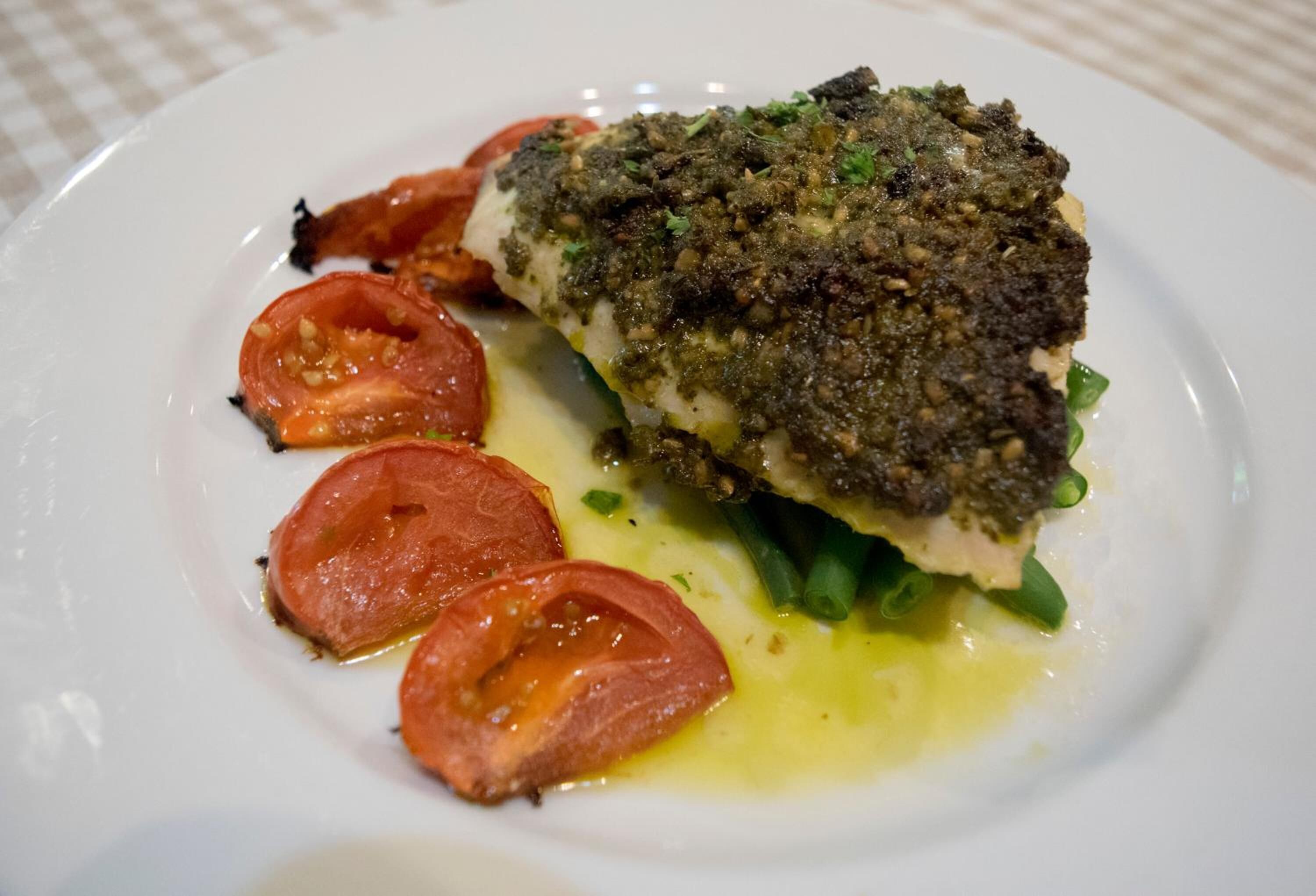 A white plate with a piece of fish topped with a green herb crust, accompanied by sliced tomatoes and green onions.