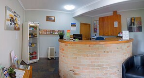 A reception area with a brick counter, a refrigerator stocked with beverages, a potted plant, and a wooden cabinet. - Rest Point Motor Inn (Glen Innes)