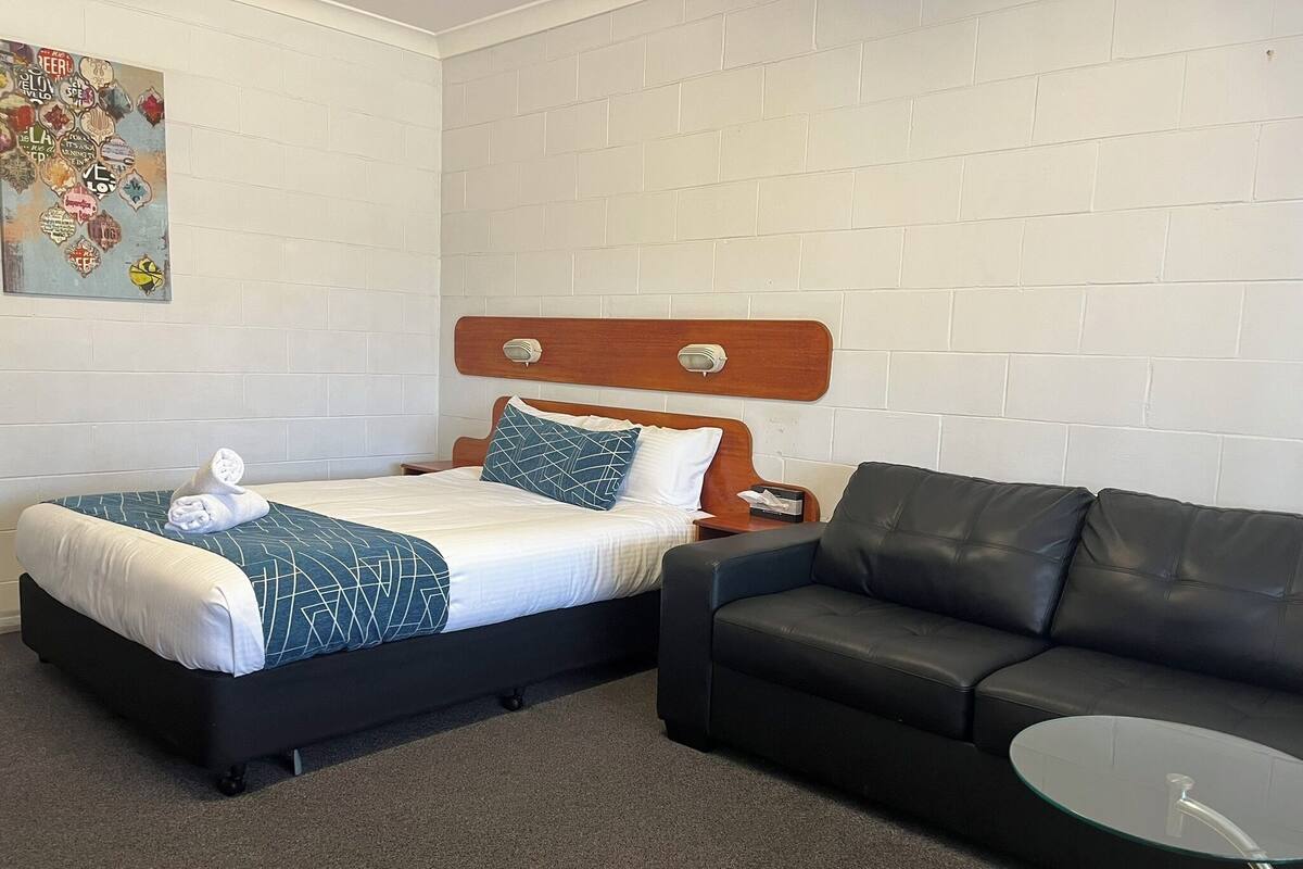A hotel room with a bed, a black leather sofa, a glass coffee table, and a wall with a decorative poster.
