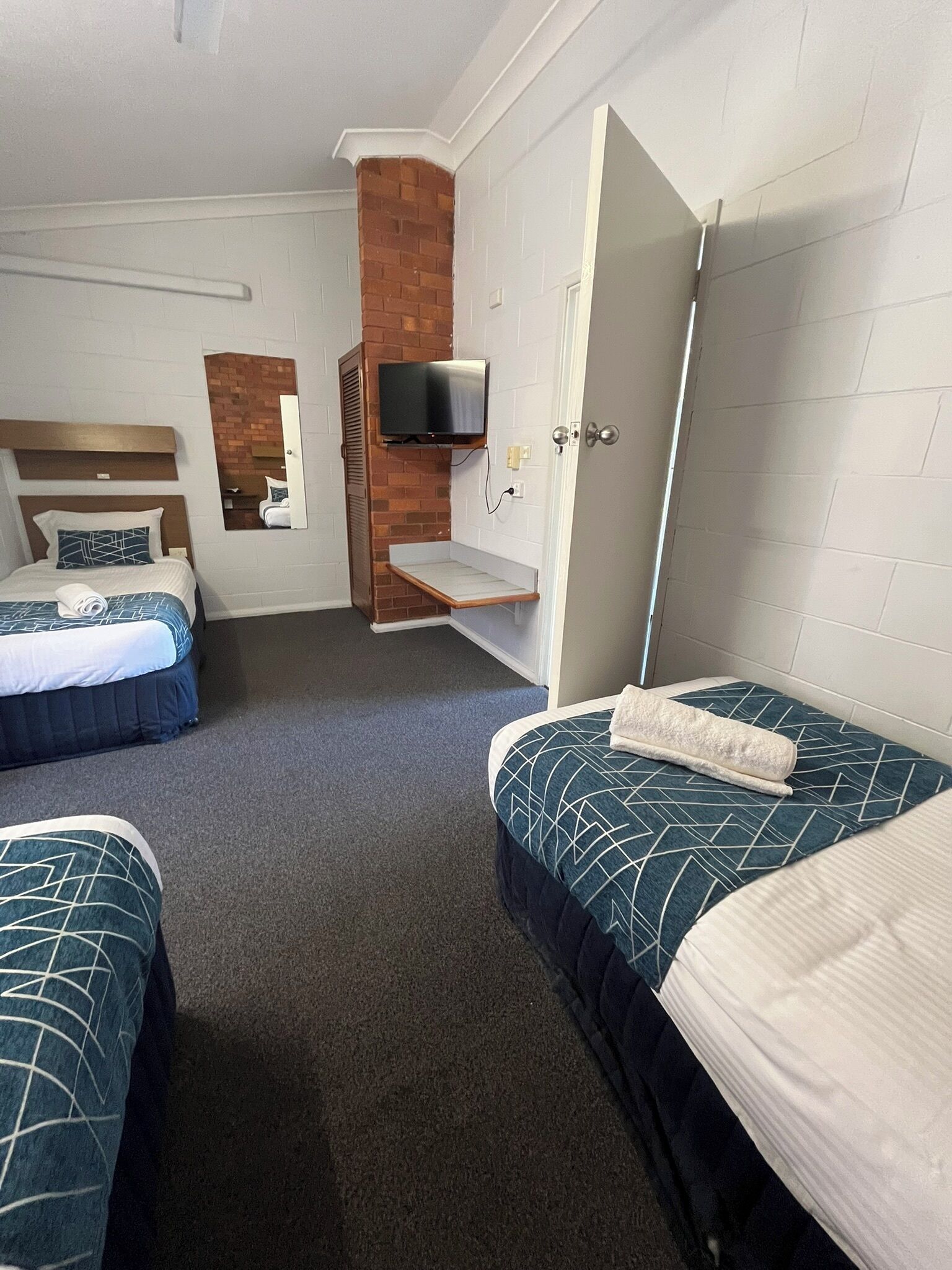 A hotel room with two beds, a TV mounted on a brick wall, a mirror, and a door leading to another room.