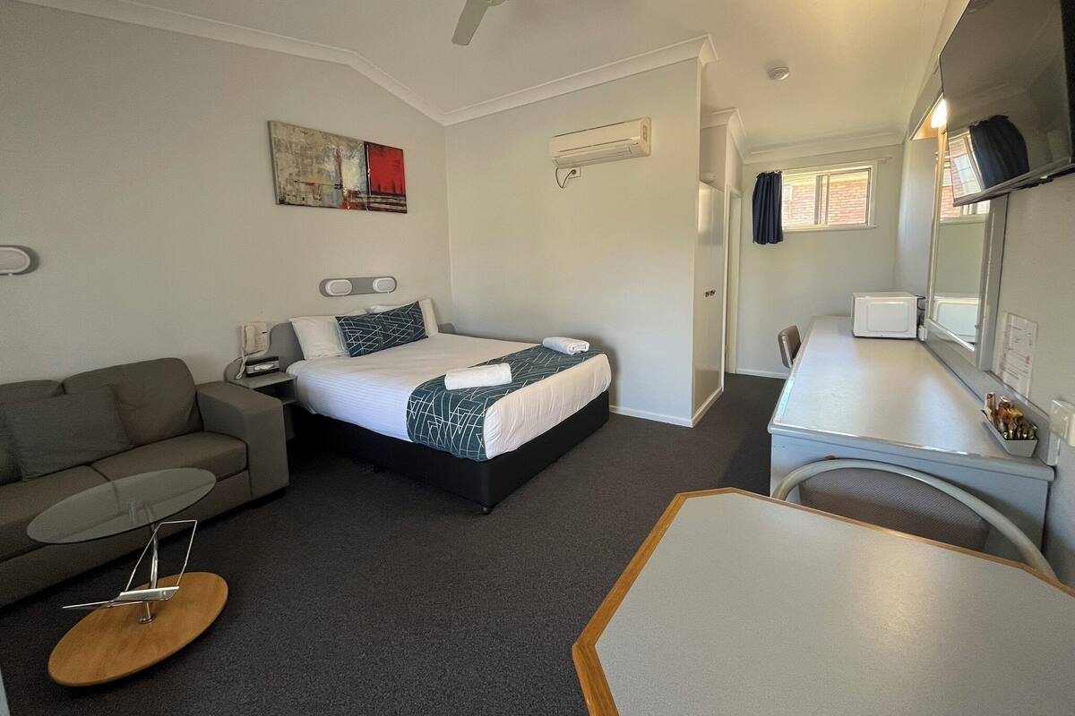 A hotel room with a bed, a sofa, a small table, a desk, a ceiling fan, an air conditioning unit, and a window.