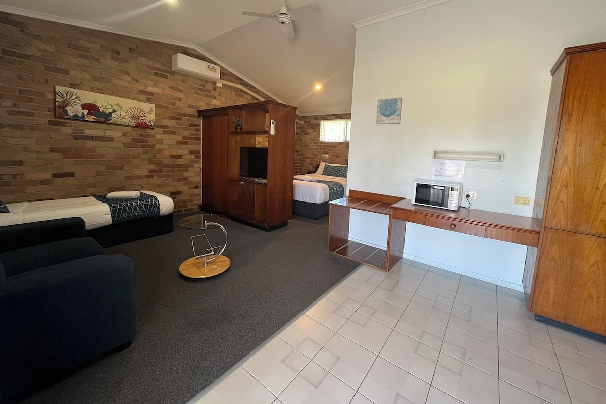A hotel room with a brick wall, a wooden wardrobe, a sofa, a bed, a small table with a microwave, and a ceiling fan.