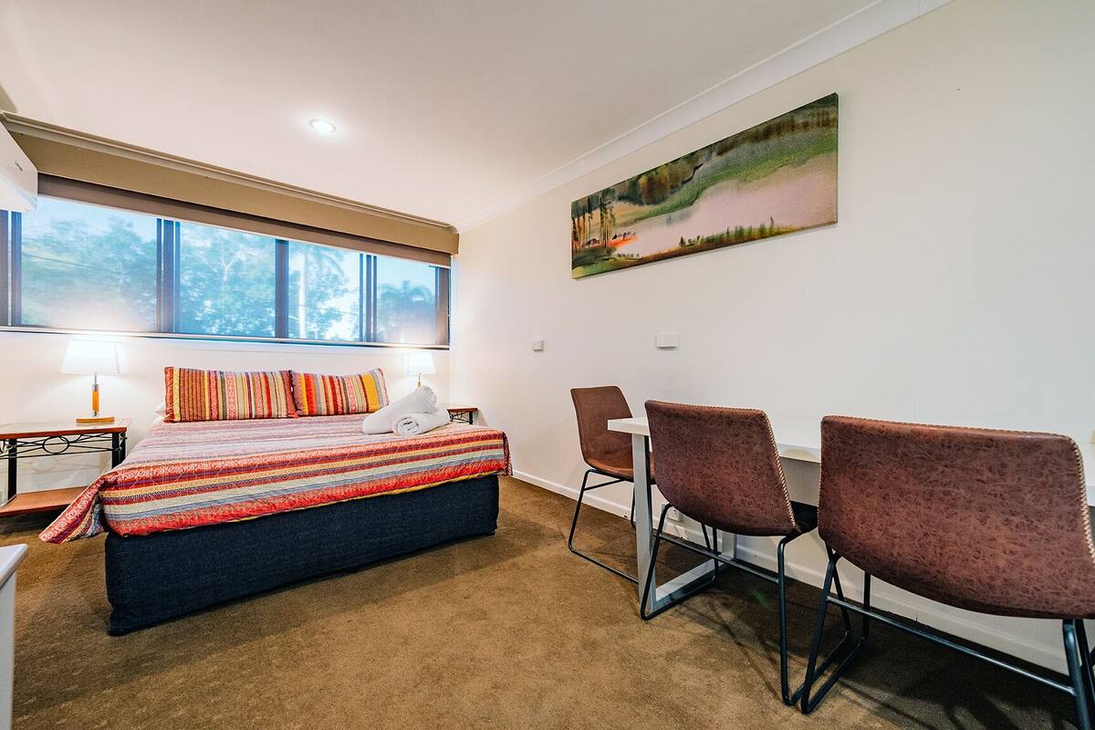 A bedroom with a bed, a desk, two chairs, a painting on the wall, and large windows.