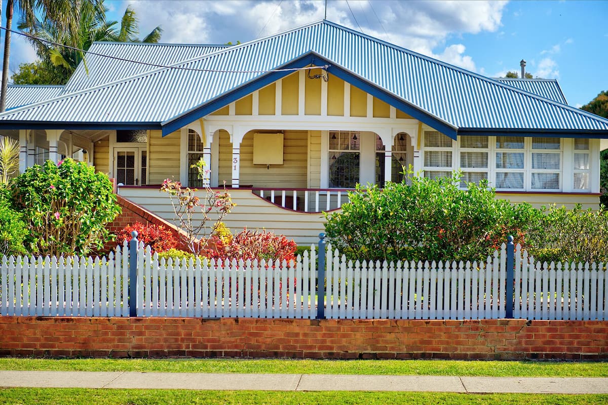 A two-story house with a blue roof, a white picket fence, and a brick wall.
