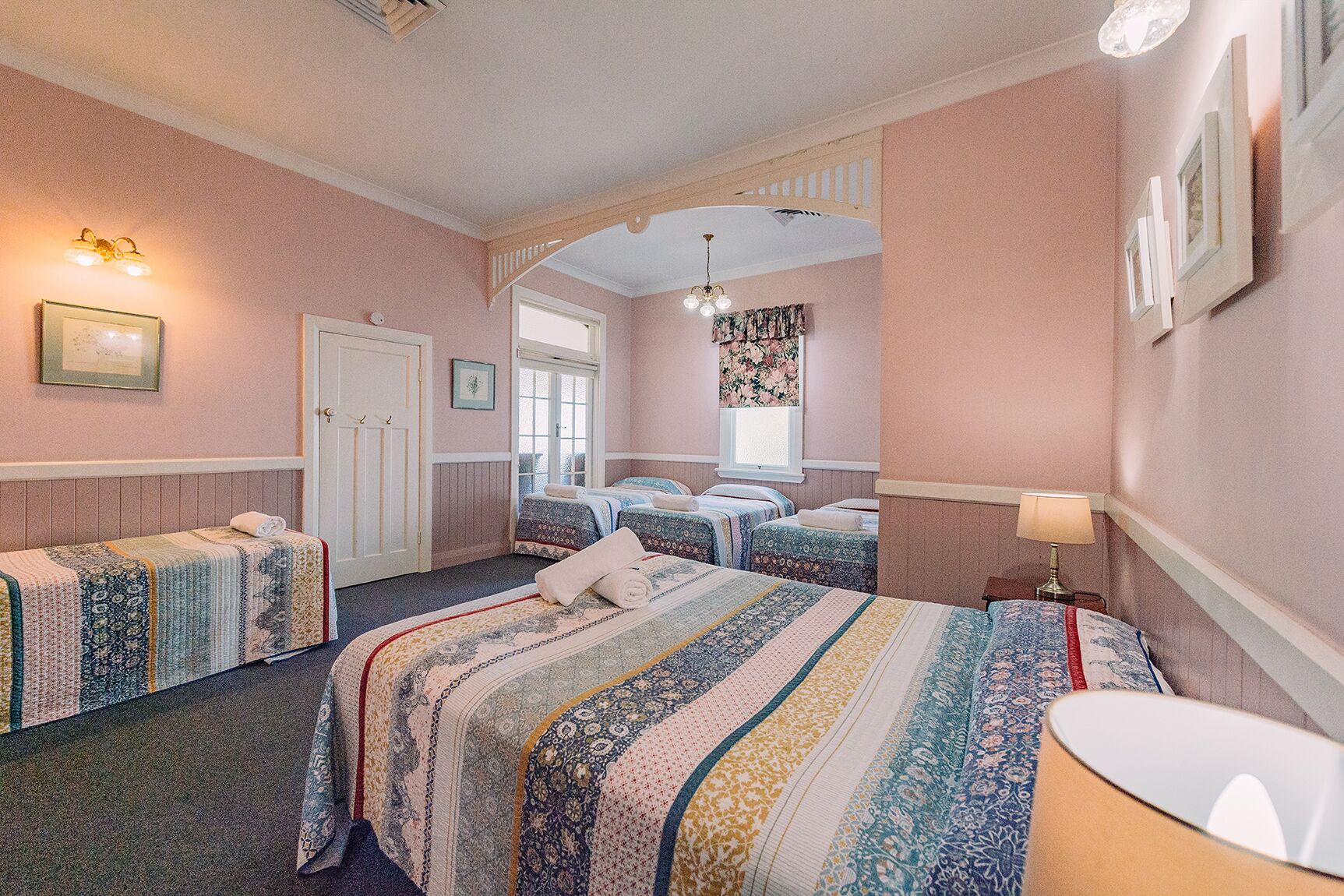 A hotel room with two single beds, a large bed, a sitting area with a chair, a desk, and a window with floral curtains.