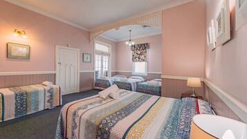A hotel room with two single beds, a large bed, a sitting area with a chair, a desk, and a window with floral curtains.