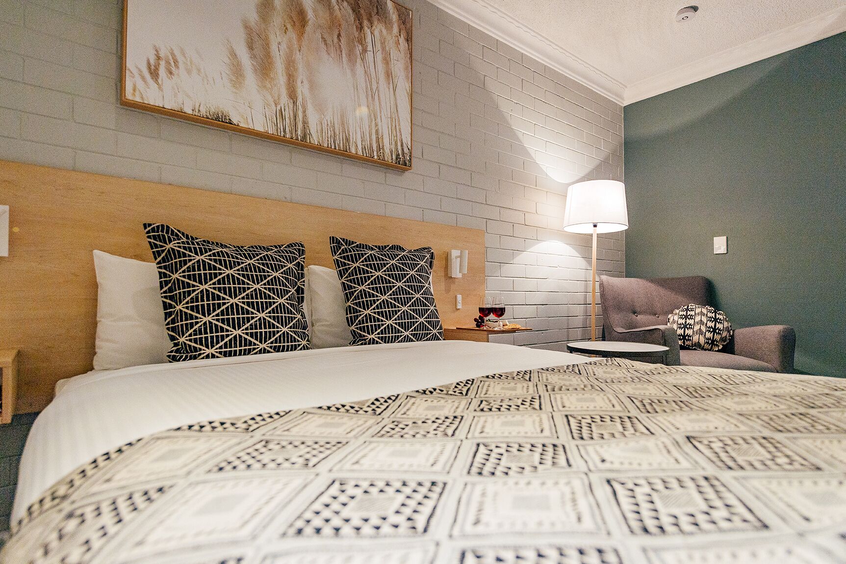 A hotel room with a large bed, a headboard with geometric pillows, a side table with a wine glass and a plate, and a lamp.