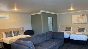 A hotel room with a grey sectional sofa, two single beds, a small table, and a door leading to another room. - Fitzroy Motor Inn Grafton (Grafton)