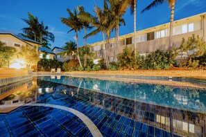 Una piscina con borde de baldosas, rodeada de palmeras y un edificio de fachada de ladrillo.