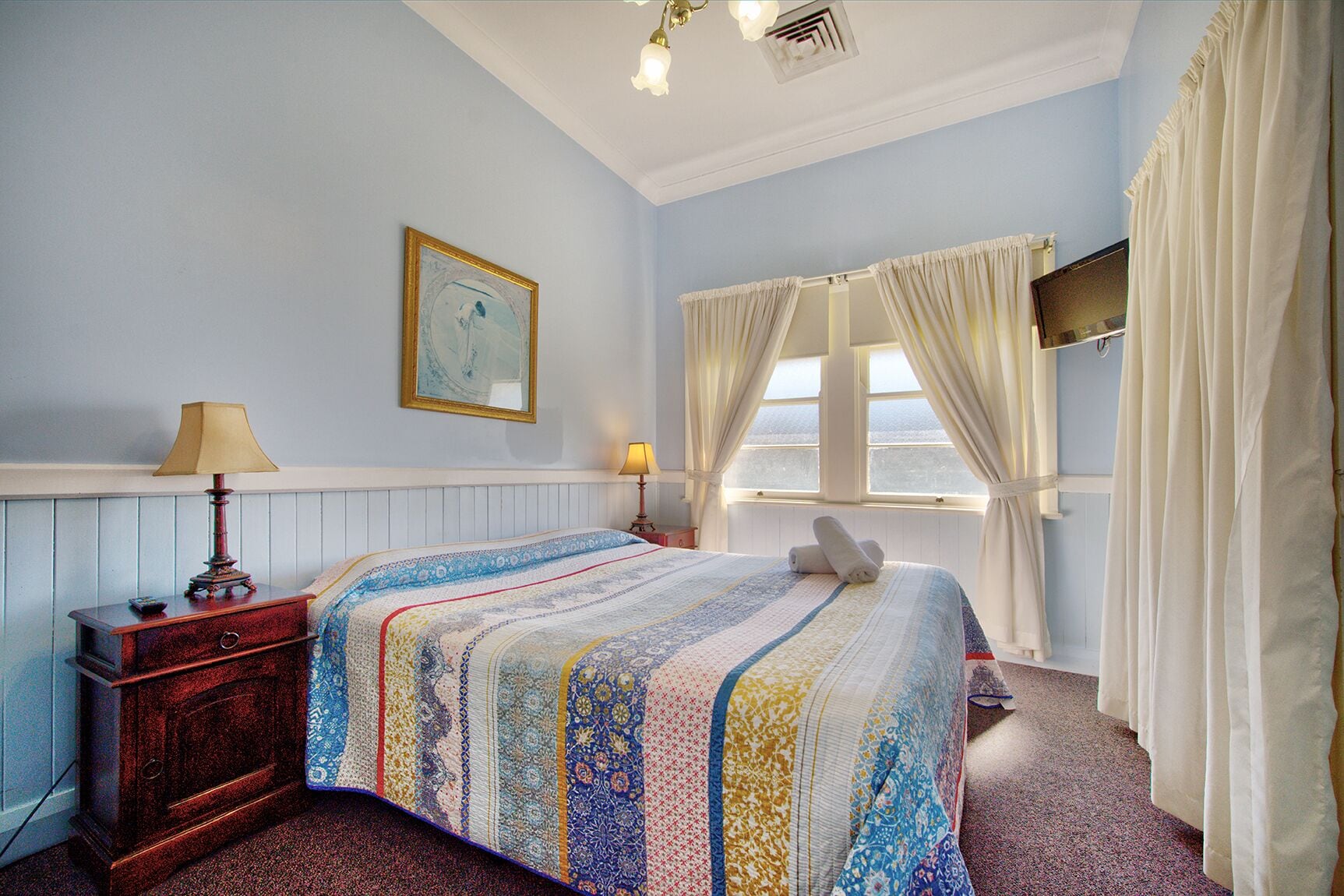 A bedroom with a bed, a nightstand, a lamp, a painting, and a window with curtains.