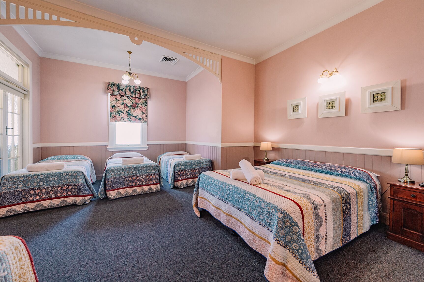 Quarto com quatro camas, um candelabro, uma janela com cortinas florais e quadros nas paredes.