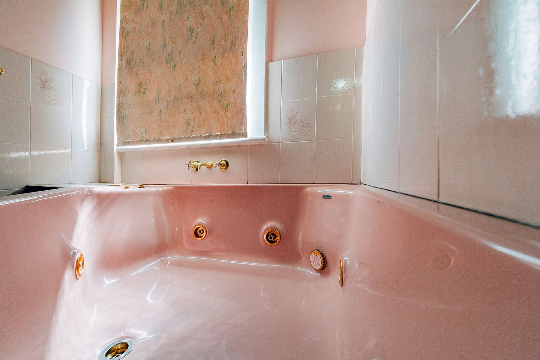 Eine rosa Badewanne mit goldfarbenen Armaturen in einem Badezimmer mit weiß gefliesten Wänden und einem Fenster mit Blumenmuster.