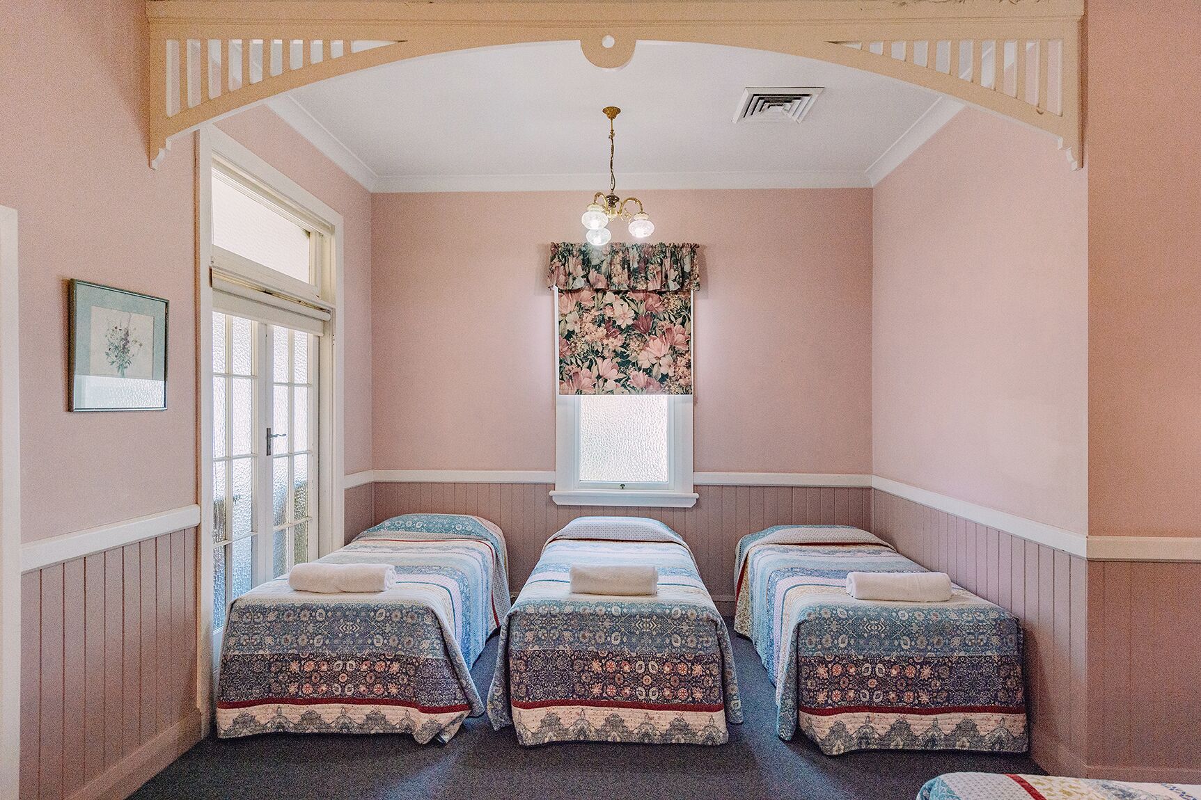 A room with three beds, a window with floral curtains, a chandelier, and a framed picture on the wall.