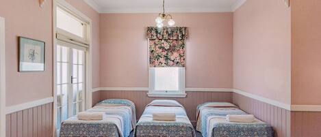A room with three beds, a window with floral curtains, a chandelier, and a framed picture on the wall.