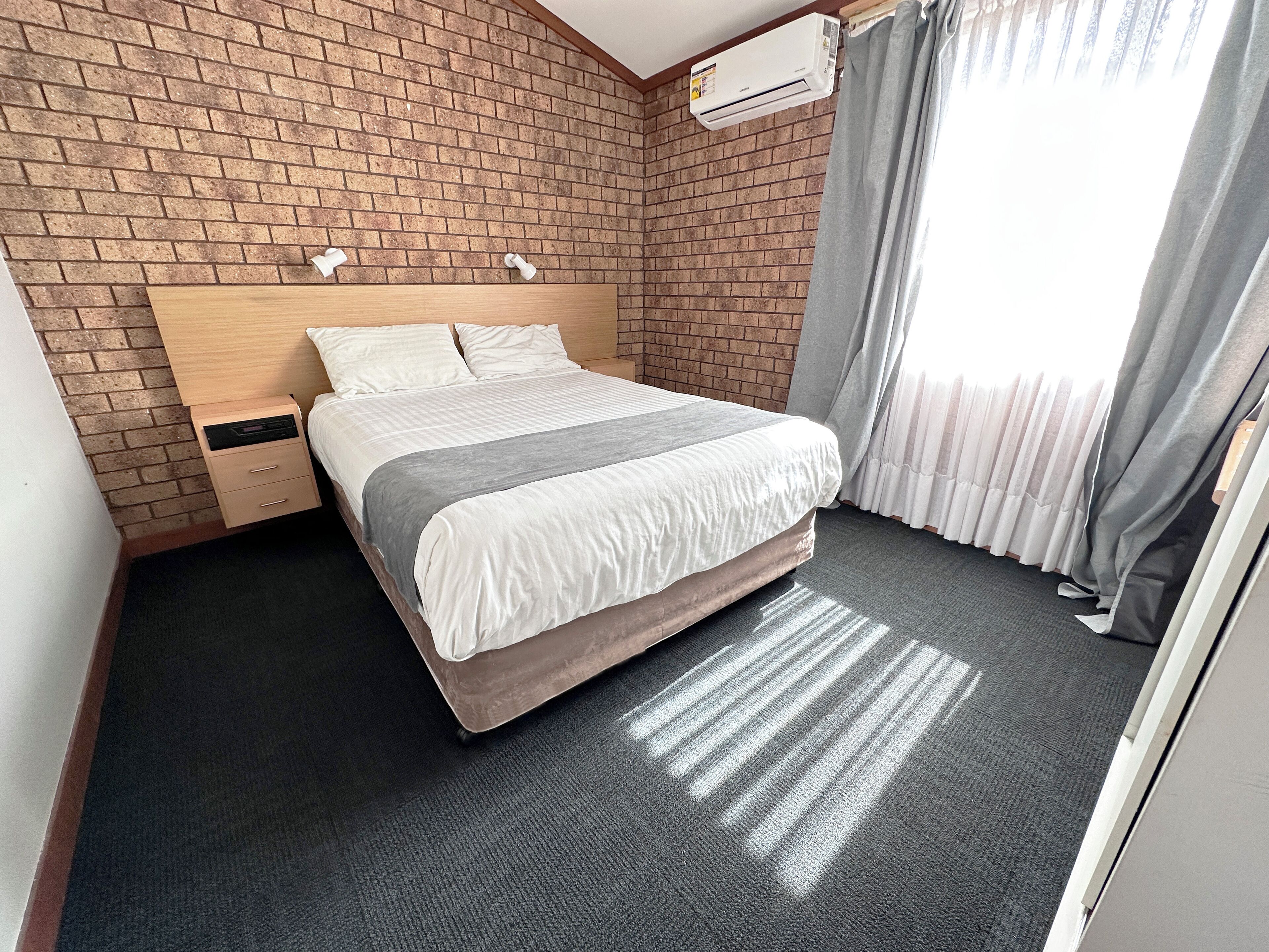 A hotel room with a brick wall, a wooden headboard, a bed with white bedding, a nightstand, and a window with curtains.