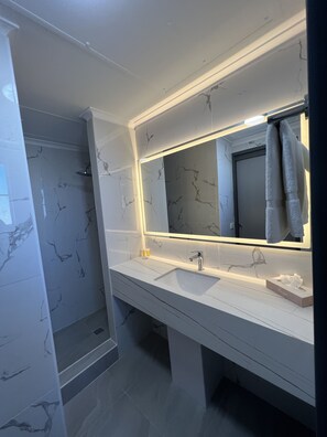 A modern bathroom with a marble backsplash, a large mirror with integrated lighting, a sink, and a shower area.
