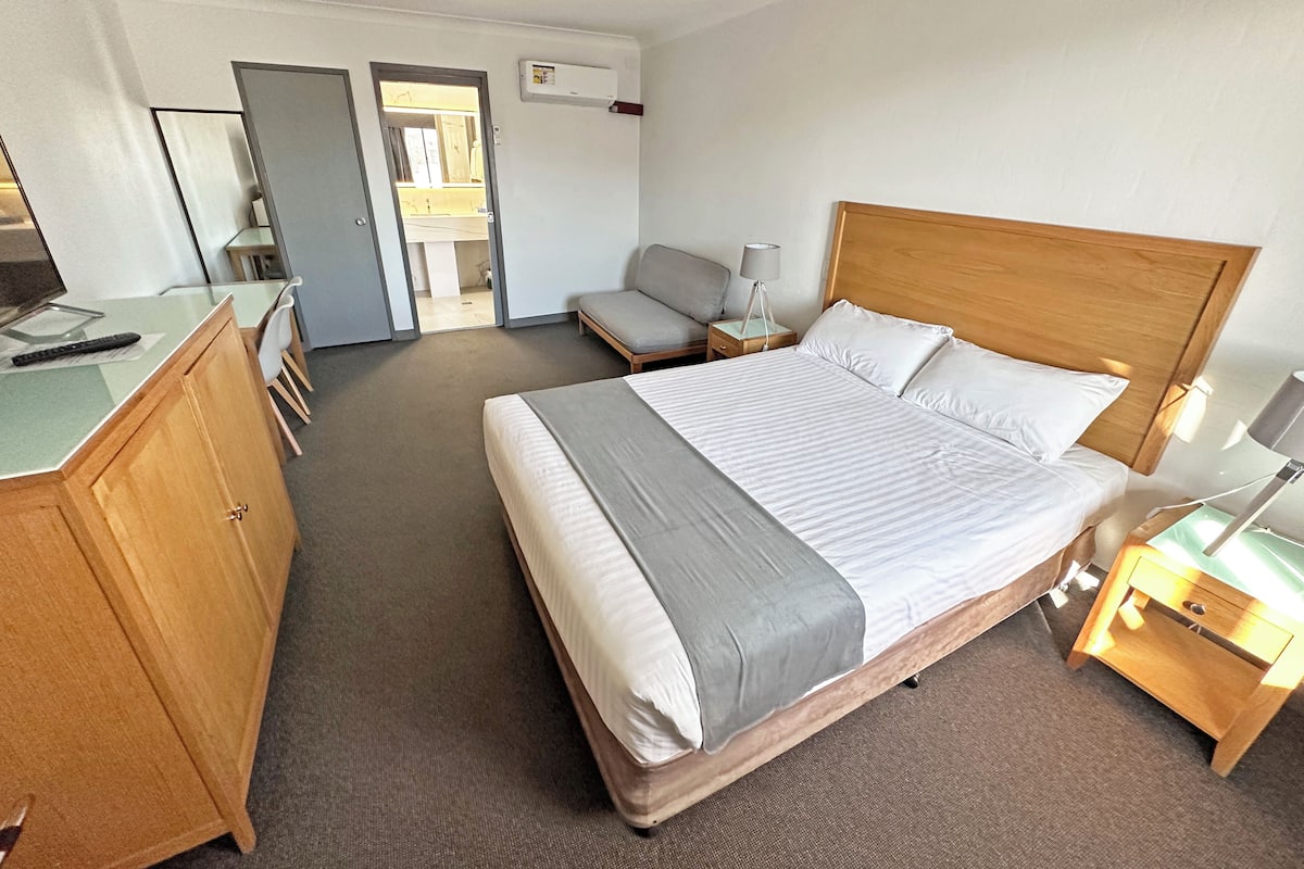 A hotel room with a bed, a desk, a chair, a nightstand, and a wardrobe.