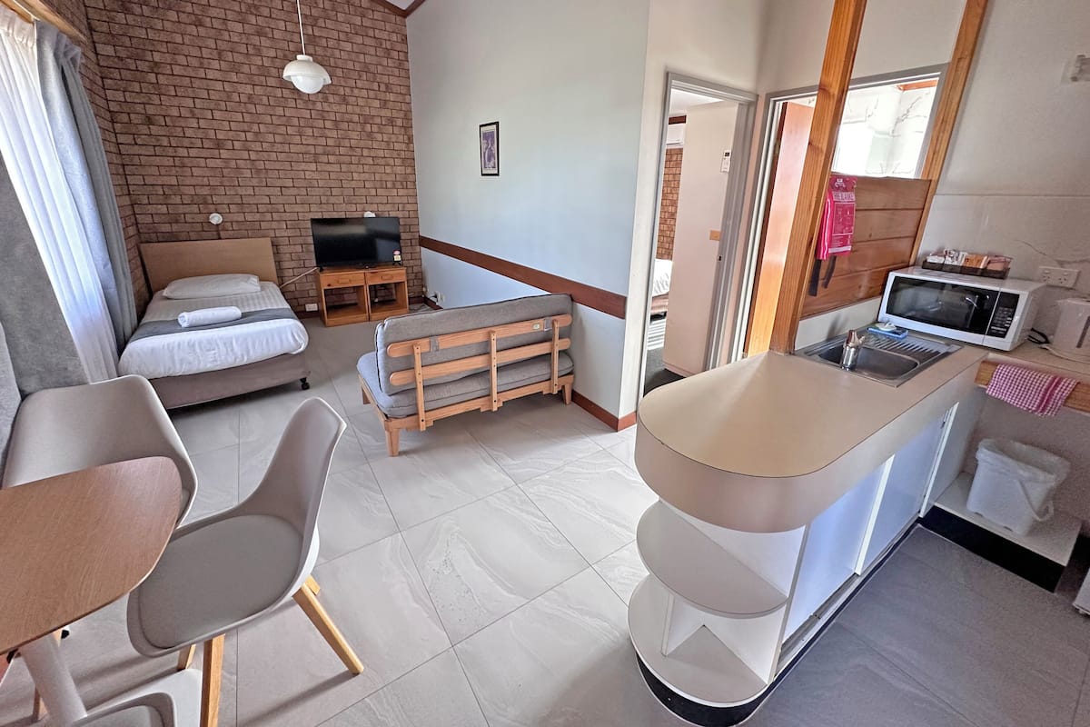 A compact hotel room with a bed, a sofa, a small dining table, a kitchenette with a microwave, and a TV on a brick wall.