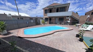 A rectangular swimming pool with a brick border, surrounded by a paved patio with potted plants. - Spa Village Motel (Moree)