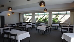 A dining area with tables and chairs, large windows, and hanging pendant lights. - Spa Village Motel (Moree)