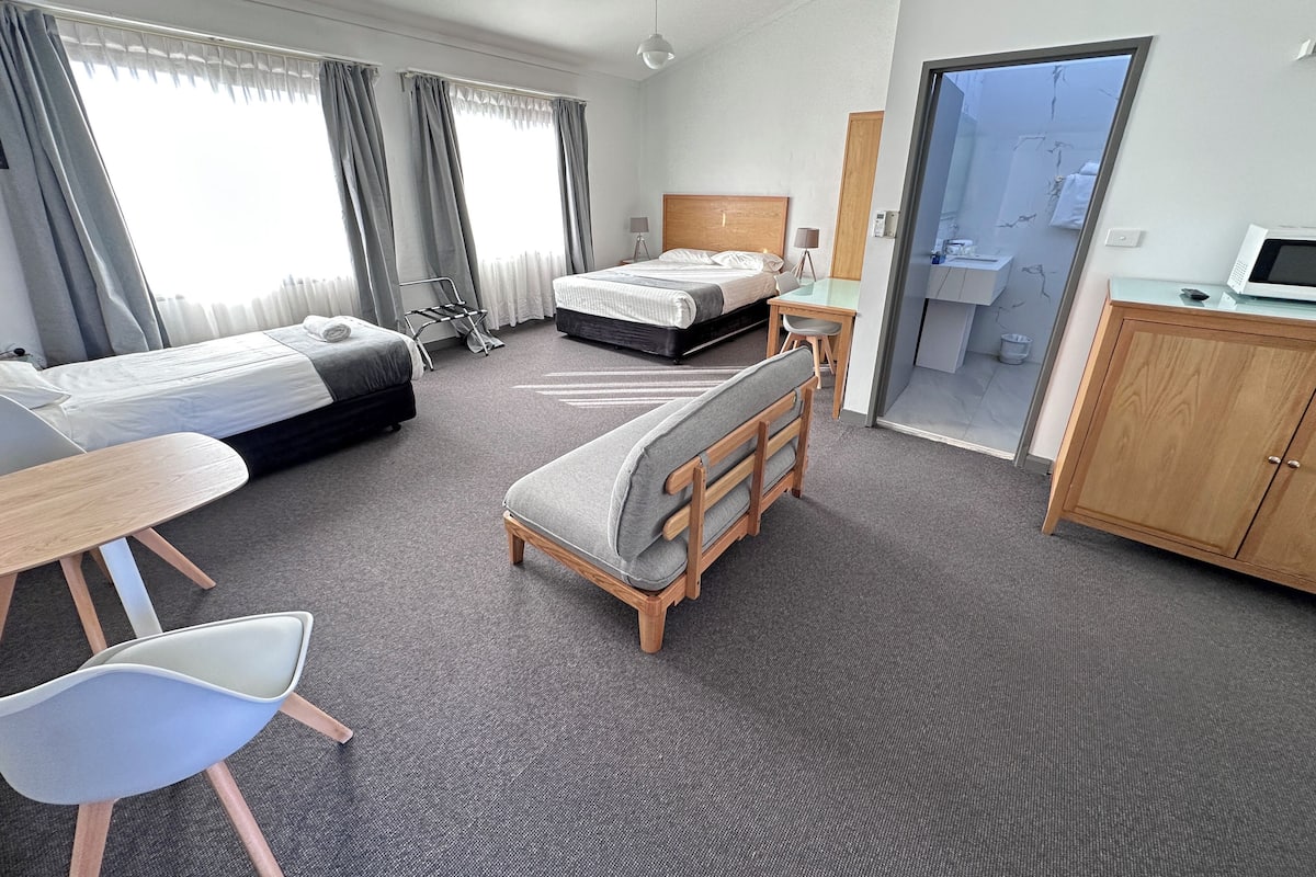 A hotel room with two beds, a desk, a chair, a wardrobe, and a bathroom visible through an open door.