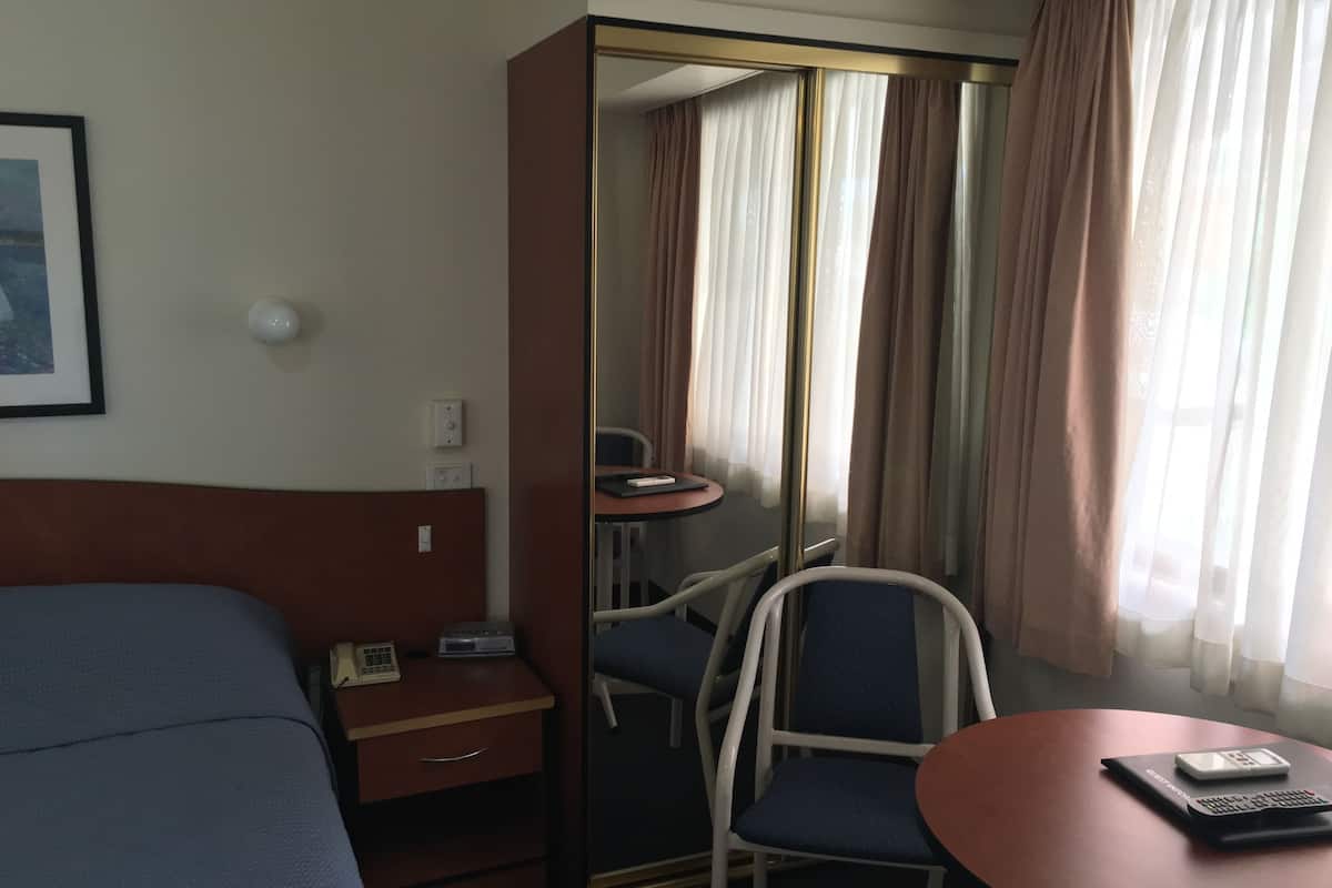 Habitación de hotel con cama, escritorio con teléfono, silla, mesita con calculadora y espejo con cortinas.