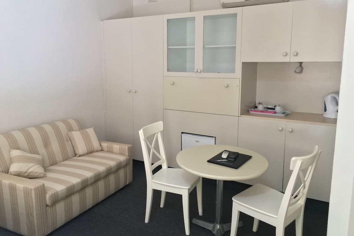 Una habitación pequeña y ordenada con un sofá a rayas, dos sillas blancas, una mesa redonda y un área de kitchenette con armarios y una cafetera.