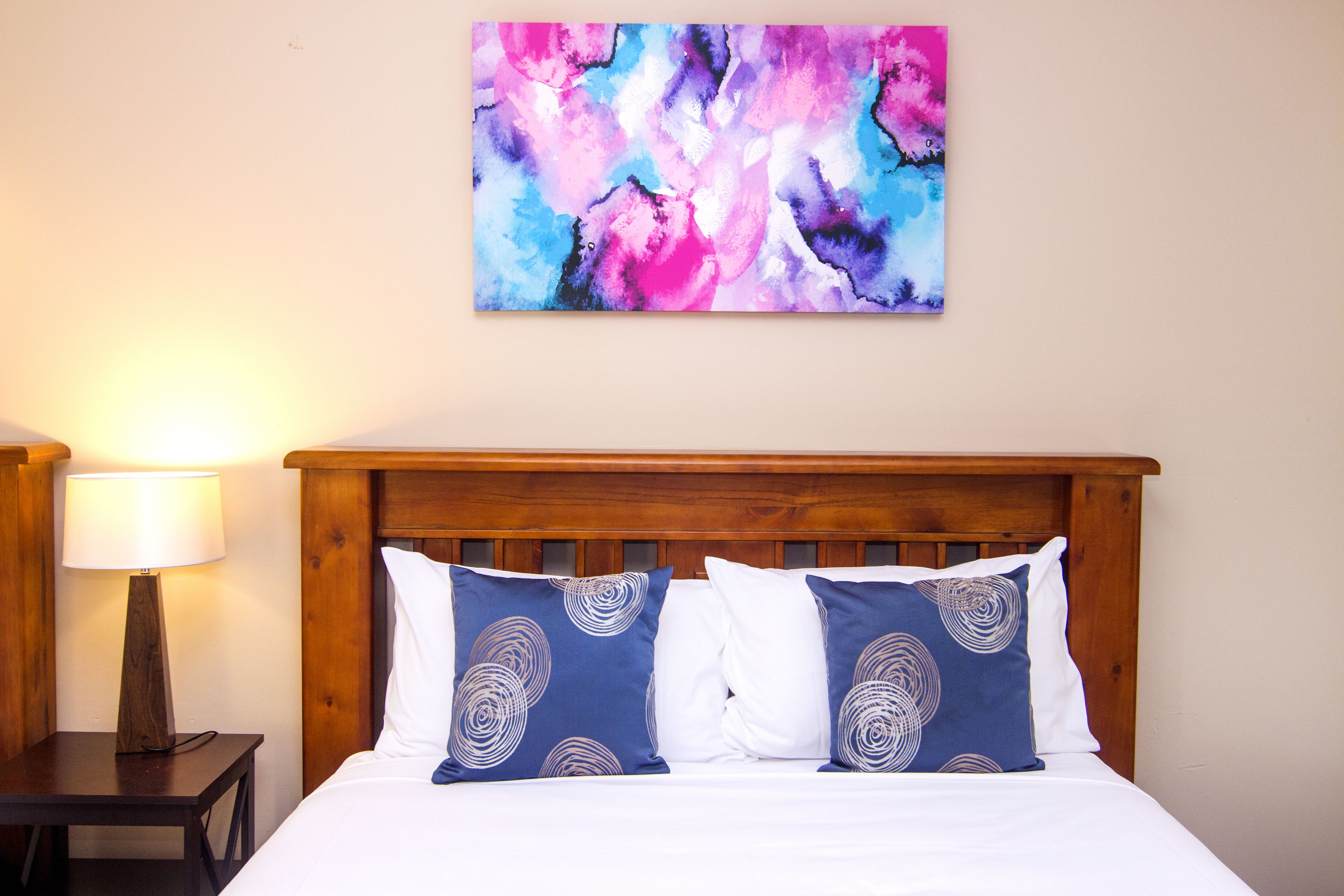A neatly made bed with two pillows, a wooden headboard, and a wall-mounted abstract painting.