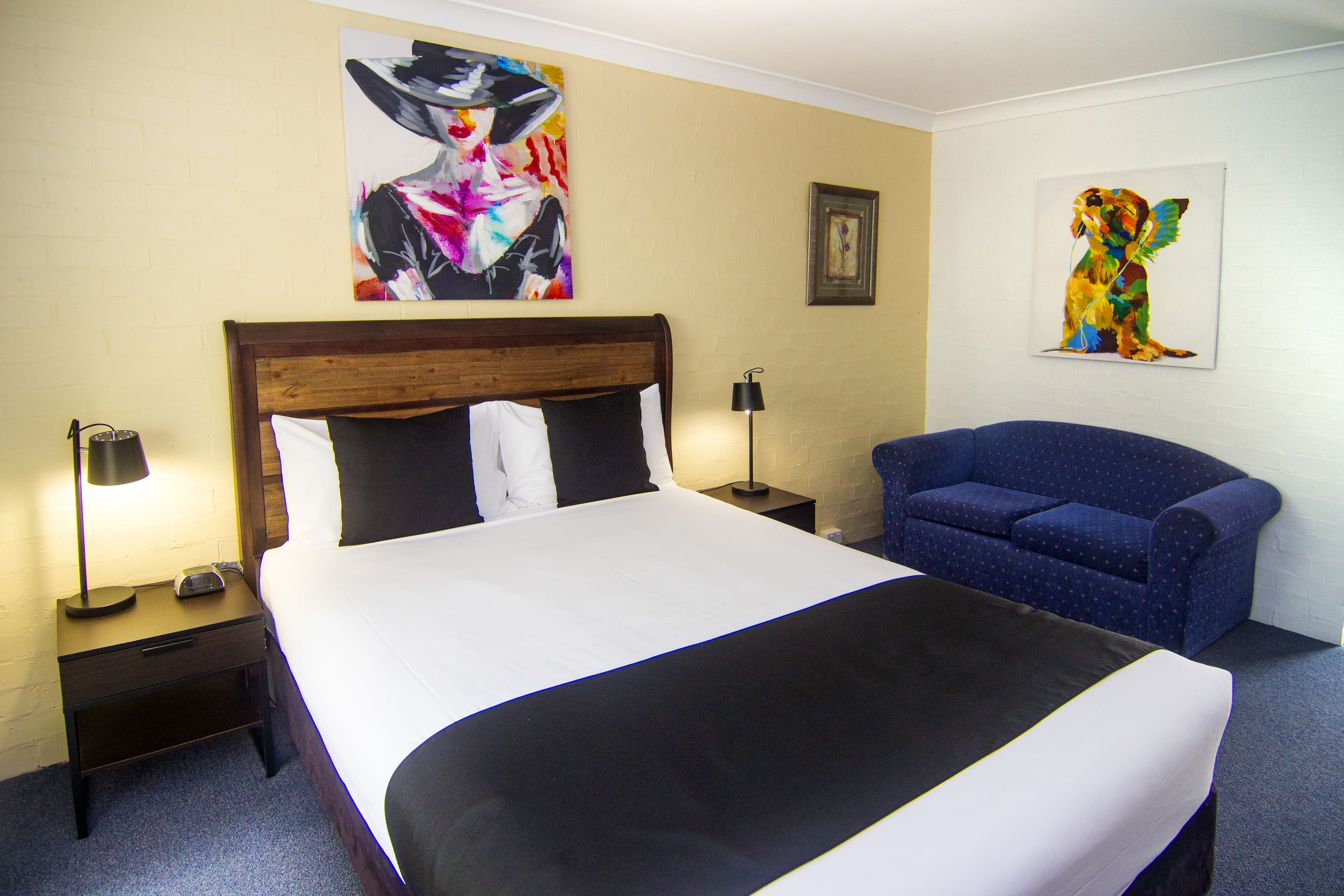 A hotel room with a large bed, a nightstand, a lamp, a blue armchair, and two pieces of wall art.
