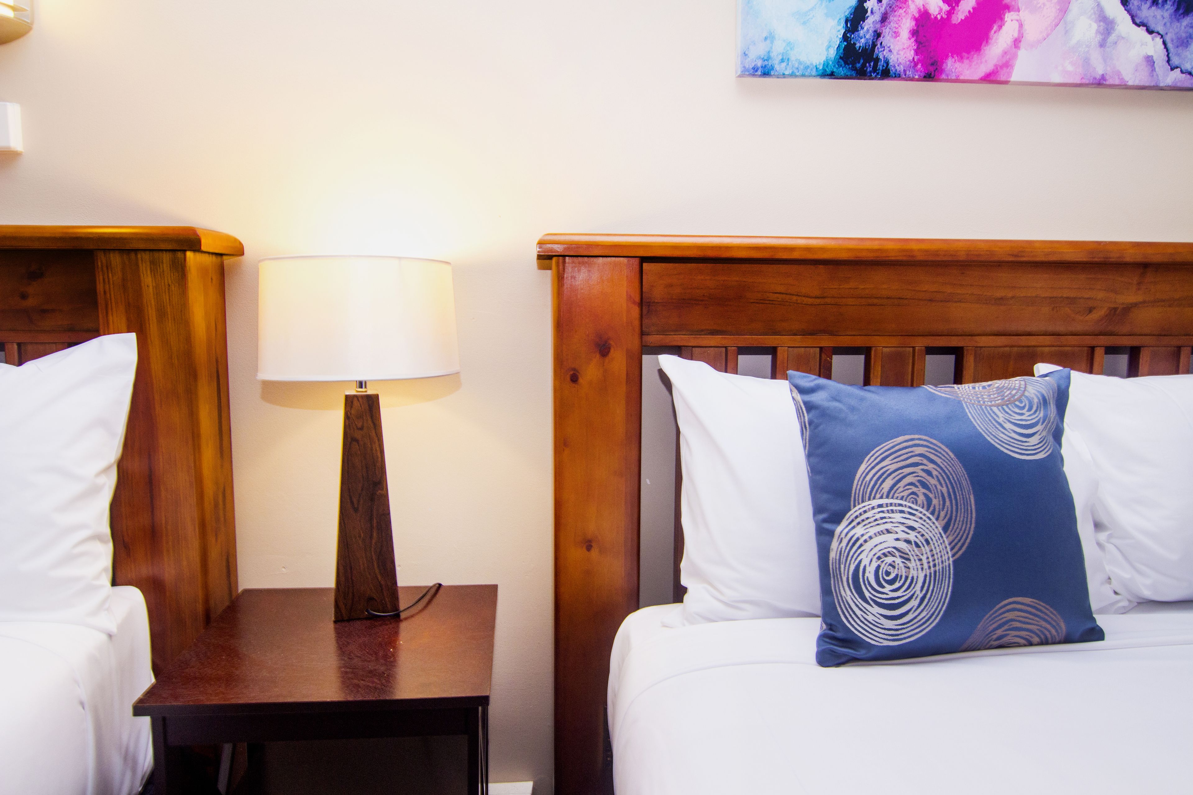A hotel room with two beds, each with a wooden headboard, a bedside table with a lamp, and a painting on the wall.