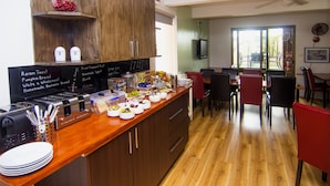 A kitchen with wooden cabinets, a countertop with various food items, a dining area with chairs and a table, and a television mounted on the wall. - Crescent Motel (Taree)