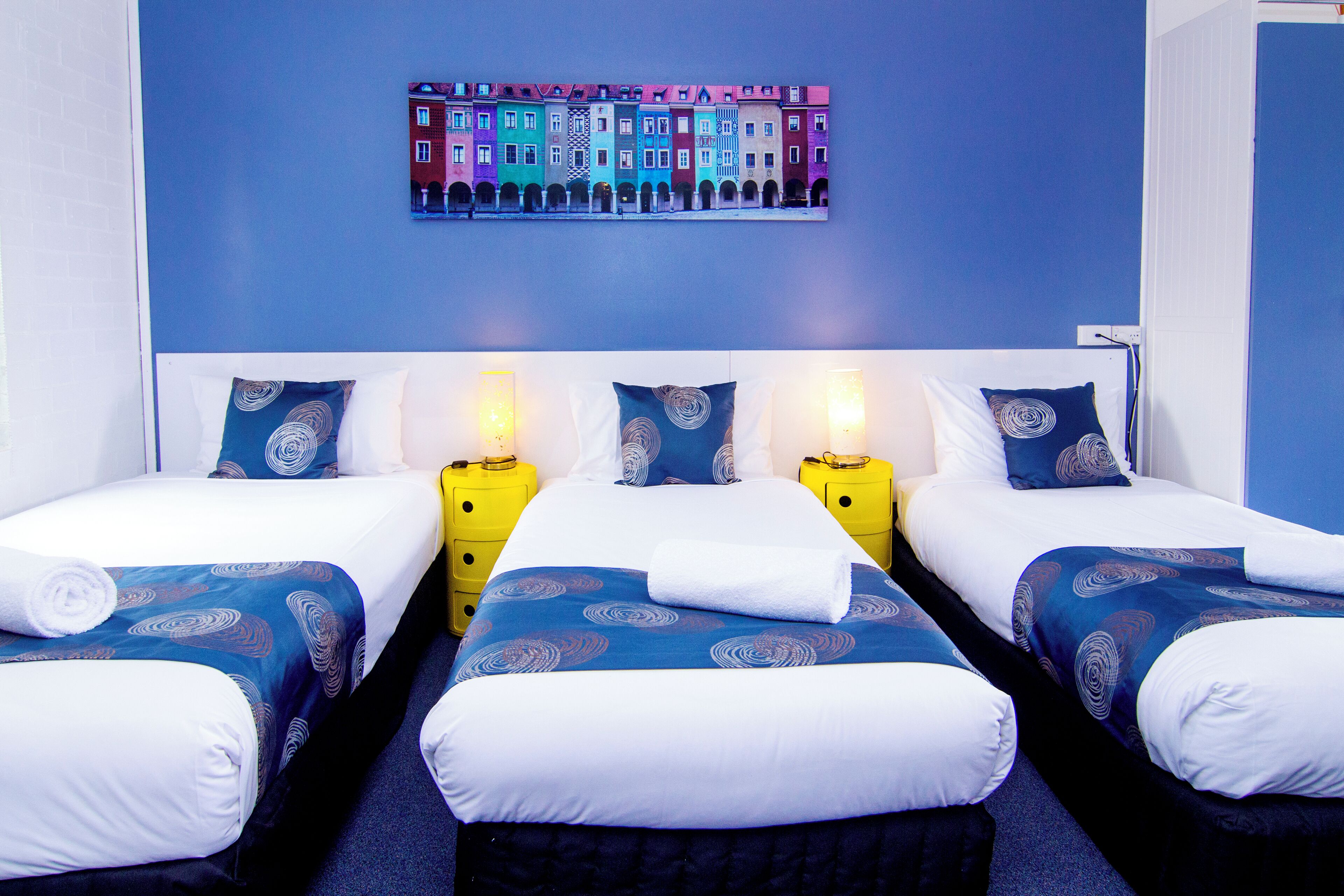 Habitación de hotel con tres camas, cada una con una colcha de diseño azul y blanco, almohadas blancas y cabecero blanco. Hay dos mesitas de noche con lámparas amarillas.