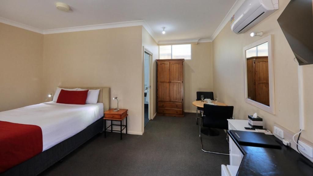 A hotel room with a bed, a desk, a chair, a wardrobe, and an air conditioning unit.