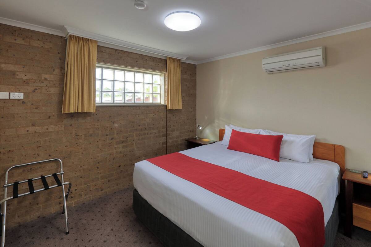 Habitación de hotel con una cama grande, una ventana con cortinas, una mesita de noche, una lámpara y un aire acondicionado instalado en la pared.