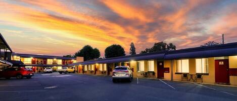 Ein Motel mit mehreren Zimmern, einem Parkplatz und einem Sonnenuntergangshimmel.