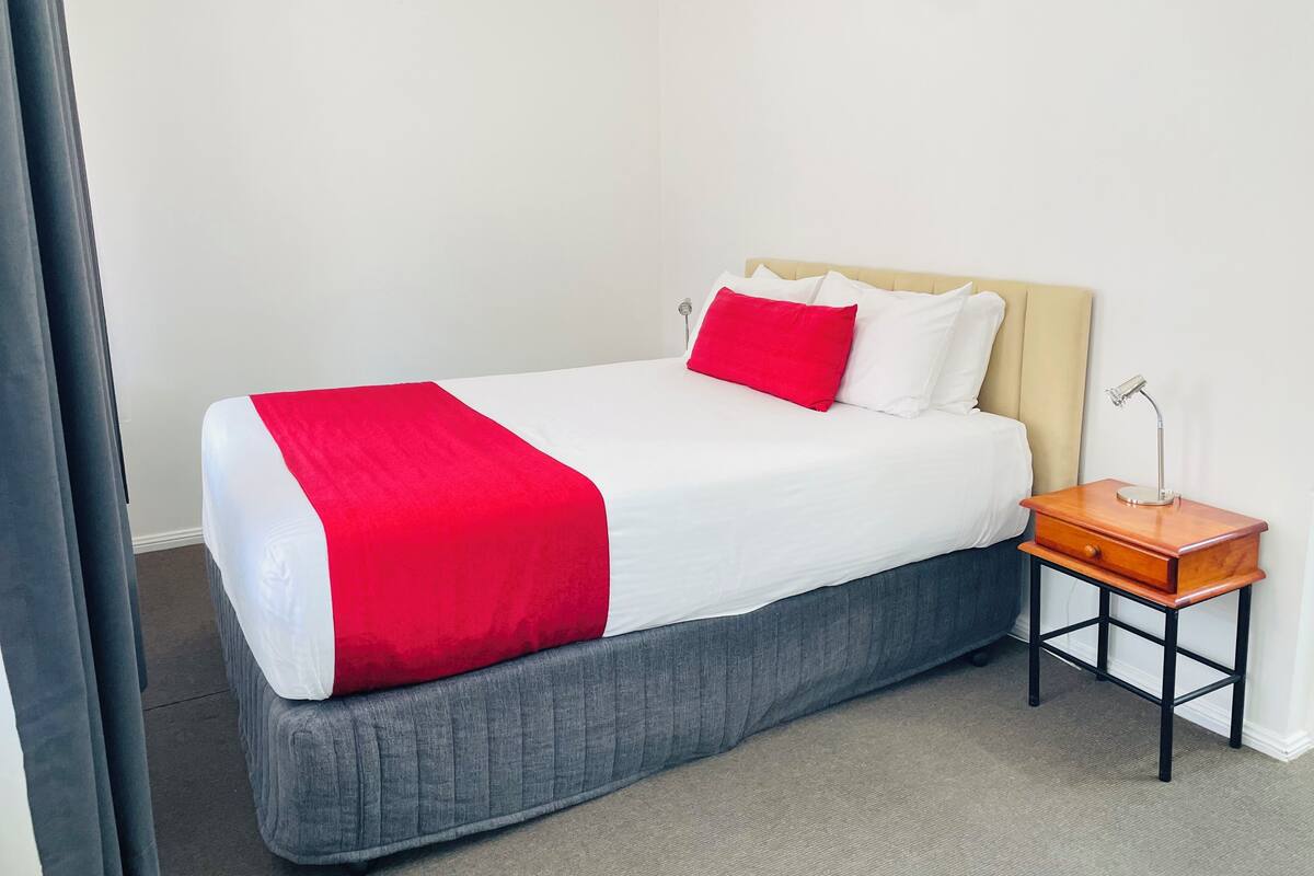 Una cama bien hecha con una colcha roja y blanca, un cabecero tapizado en gris y una mesita de noche de madera con una lámpara.