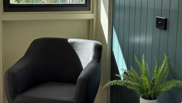 A room with a window, a chair, a bed, a wall-mounted light, and a potted plant. - The Heritage Darwin (Darwin)